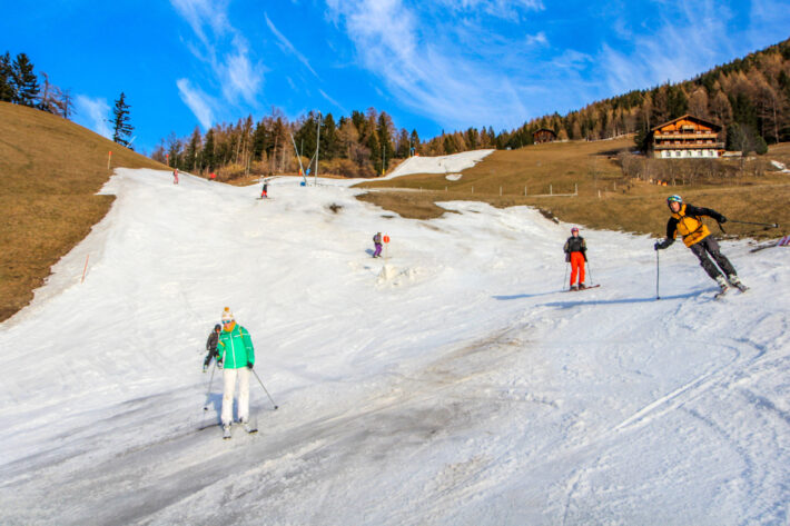 Skidåkare i vit pist med slaskig snö, omgiven av gulgröna ängar utan snö.